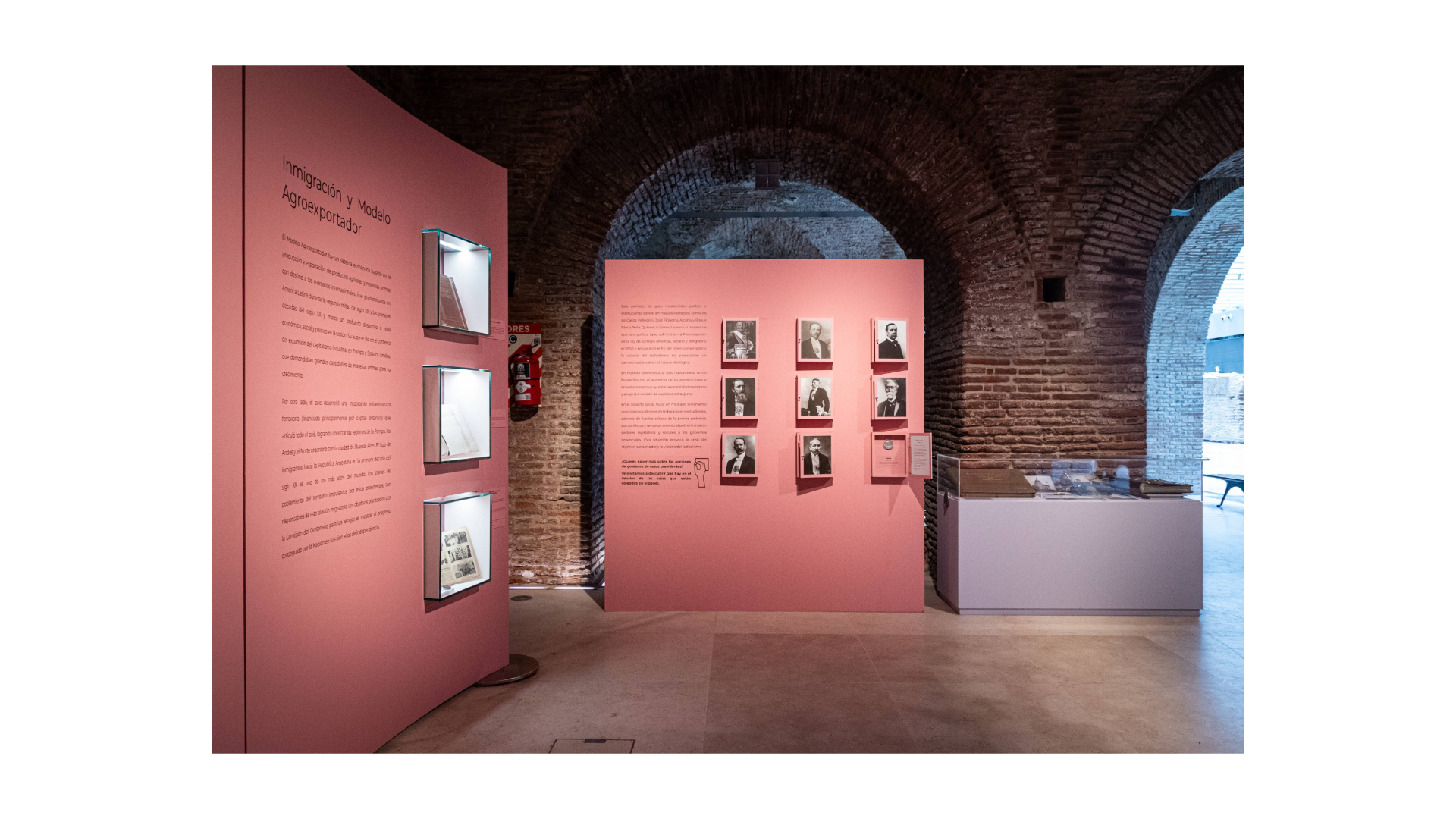 Vista de los paneles rosados de la exposición "Institucionalización del poder político". Se ven dos paneles que llevar objetos colgados. Al fondo, una vitrina apoyada sobre una pared de ladrillos.