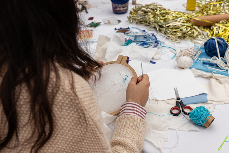 Un niña se encuentra de espaldas a la cámara. Sostiene un bastidor de bordado y está bordando con hilo celeste. Tiene cabello largo y castaño. Viste un sweater rosado. Detrás se ve la mesa con hilos, ovillos de lana, tijeras.