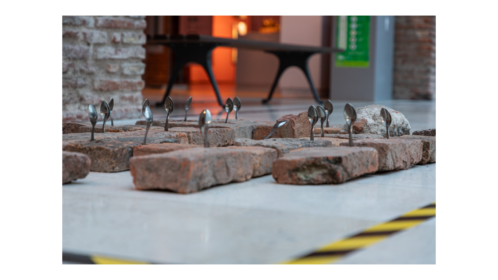 Fotografía a color de una instalación de Federico Fisher. La misma consta de ladrillos de barro colocados sobre el piso y entre ellos se apoyan cucharas pequeñas metálicas,