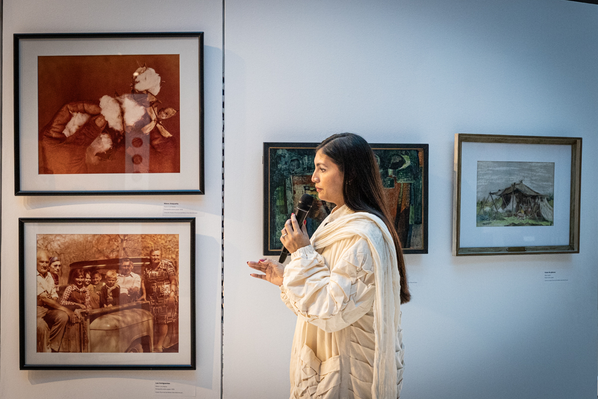 Fotografía color. En el centro se observa a Gabriela Zalazar con un micrófono en la mano. Se encuentra de perfil y por detrás se ven las obras de la exposición.