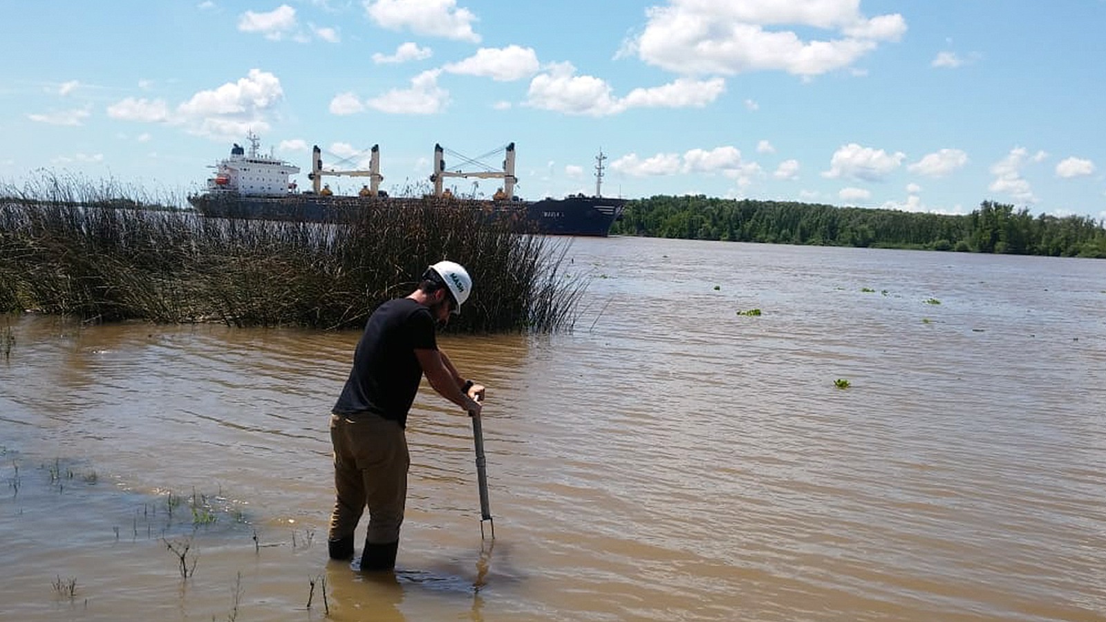 cnea científico varon con casco blanco inserta un instrumento en el agua para investigar sus propiedades con un barco de fondo