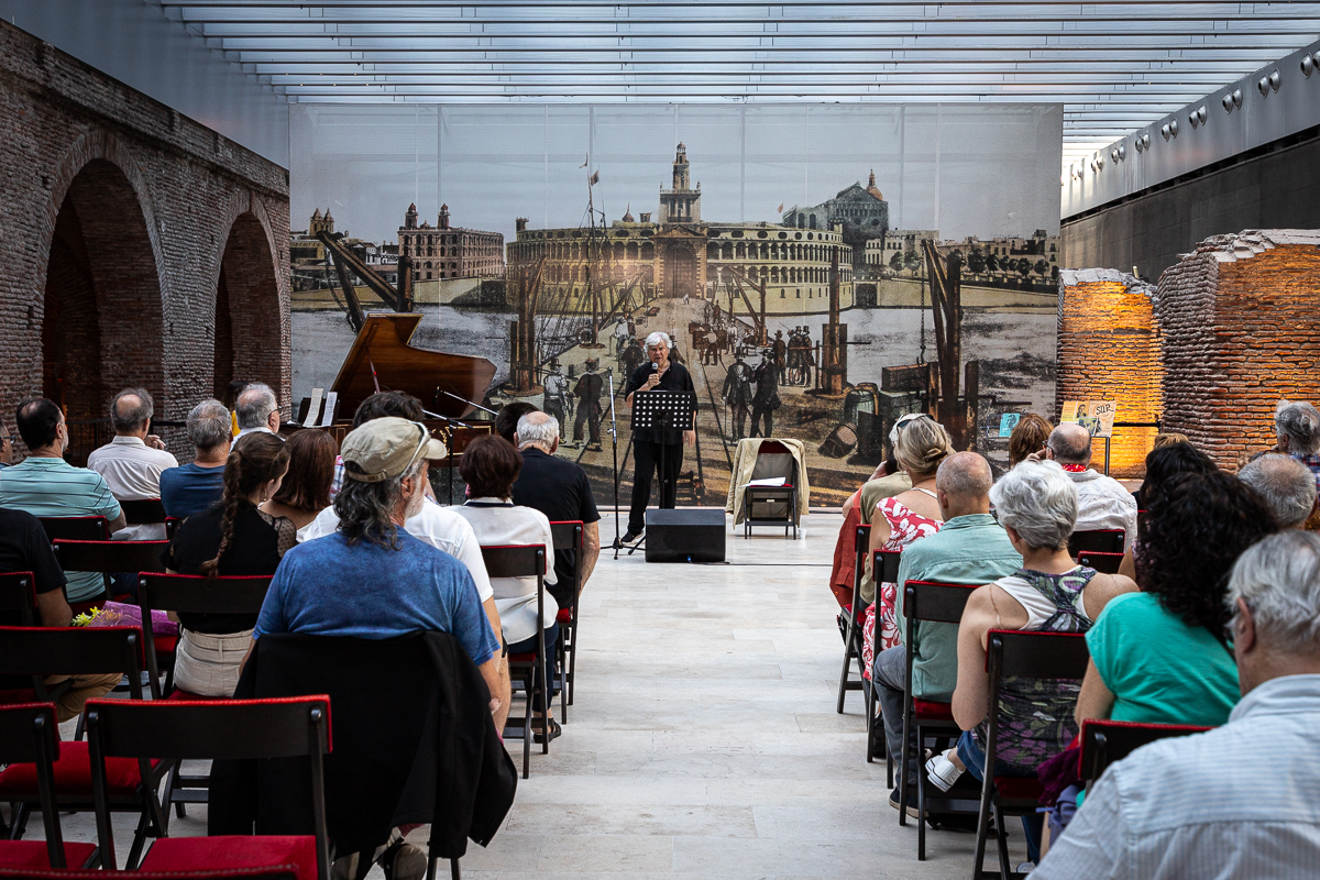 Fotografía de un evento en un espacio interior donde se ven personas sentadas en filas de sillas, mirando hacia un escenario. En el escenario hay una persina de pie frente a un micrófono cantando. Detrás de la persona hay una gigantografía de una escena histórica de una ciudad con un edificio antiguo y un muelle. A la derecha de la imagen, se pueden ver estructuras de ladrillo que son parte del diseño interior del lugar. La audiencia está compuesta por personas de diversas edades.