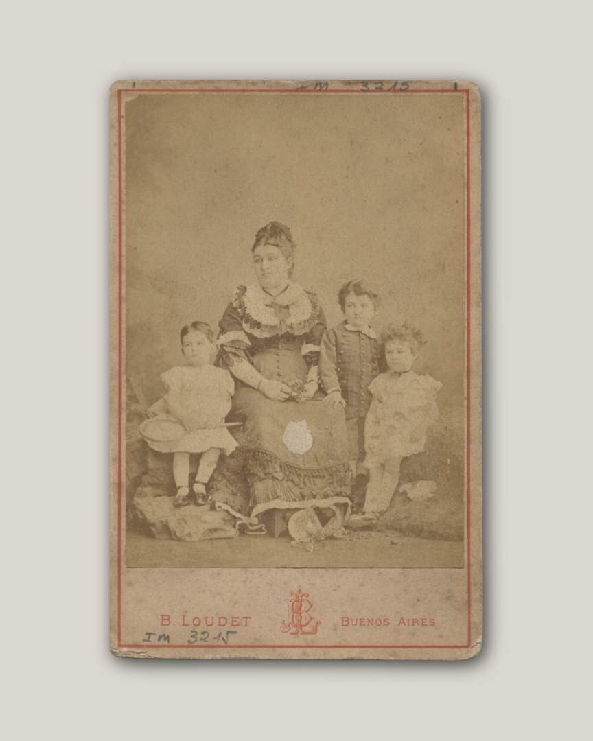 Fotografía de un grupo de personas. En el centro una mujer con vestido oscuro y cabello recogido. A la izquierda hay un niño de dos años vestido de color claro, sostiene una raqueta en sus manos. A la derecha de la mujer hay dos niños: un varón más alto vestido de oscuro y por delante una niña vestida de blanco, sentada. Debajo se lee "B. Loudet. Buenos Aires". La fotografía es sepia y blanco.
