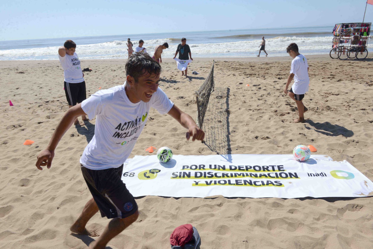 Jóvenes juegan al fútbol-tenis en la playa con una bandera de la campaña INADI en la arena con la leyenda "Por un deporte sin discriminación y violencia"