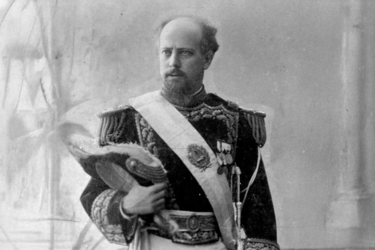 Fragmento de un retrato oficial de Julio Argentino Roca. La imagen está en blanco y negro. El expresidente se encuentra posando de frente, sobre un fondo claro. Lleva uniforme militar y banda presidencial. En su mano derecho lleva su cubrecabezas estilo bicornio.