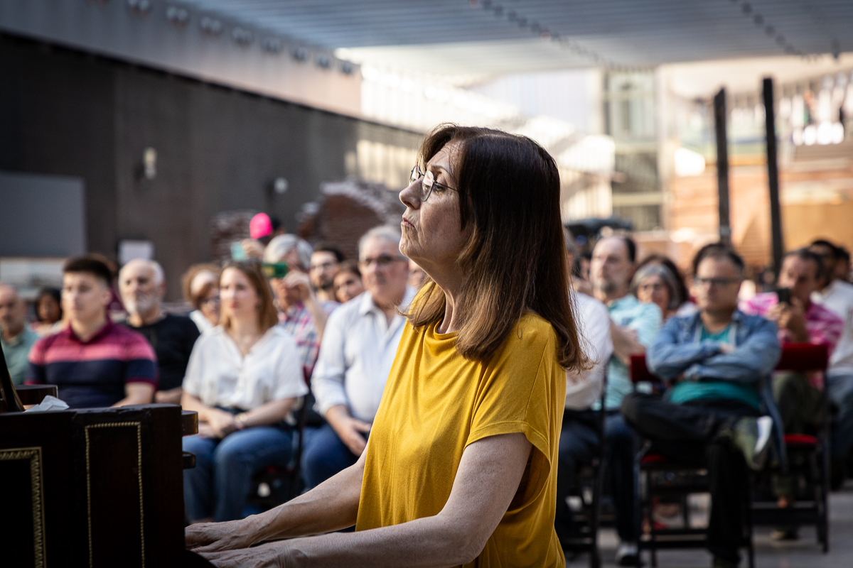La imagen muestra una mujer de perfil con cabello castaño y gafas, vistiedo una camiseta amarilla. Está sentada frente a un piano, que puede parcialmente en el lado izquierdo de la imagen. Detrás de ella hay un grupo de personas sentadas mirando hacia el frente.