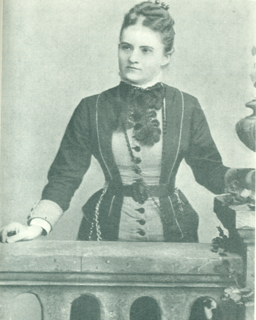 La imagen muestra una fotografía antigua en blanco y negro de una mujer de pie. Está vestida con un vestido de estilo victoriano, que tiene detalles oscuros y claros, con lazo en el cuello. Su cabello está recogido. Ella está apoyada sobre una barandilla o un mueble, su mano derecha descansando sobre él. 