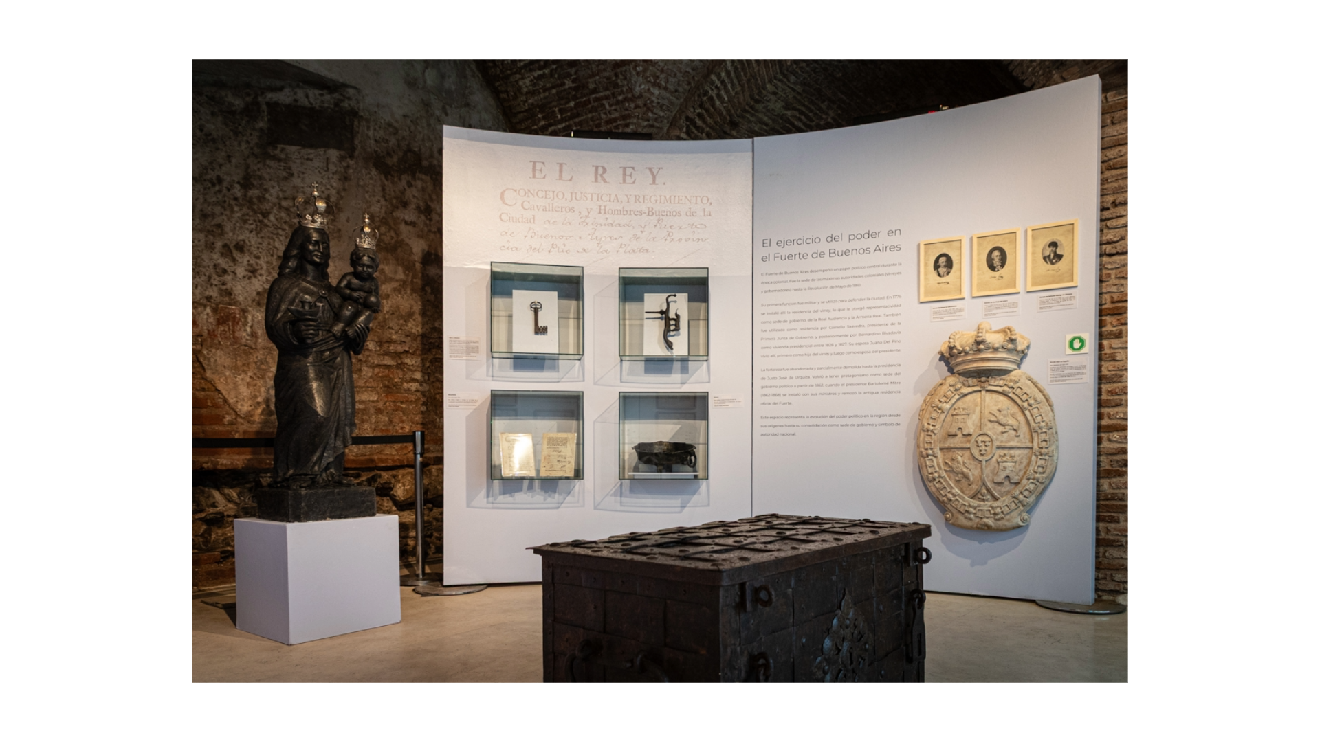Vista de la exposición "El Fuerte". A la izquierda se encuentra la estatua de la Virgen del Buen Ayre. A la derecha un panel cruvo que tiene cuatro nichos con distintas piezas. En el centro hay un arcón de hierro y a la derecha, un escudo de piedra. Por encima del escudo hay tres retratos en blanco y negro. Los nichos, el escudo y los retratos se encuentran colgados de un panel gris, que tiene un texto.