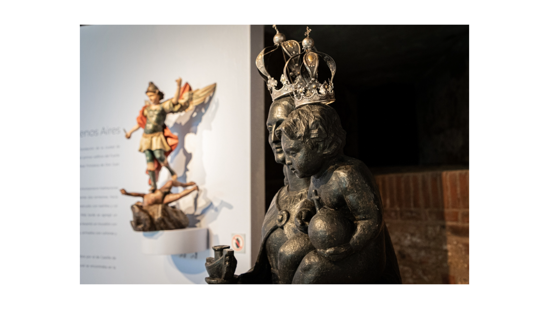 Imagen de dos esculturas. Al fondo se observa una escultura de madera policromada que representa a San Miguel Arcángel. Se encuentra sobre un estante y de fondo tiene un panel gris. A la derecha y en primer plano hay un fragmento de una escultura de yeso pintado de negro. Esta pieza representa a la Virgen del Buen Ayre. En la cabeza de la estatuta se encuentra una corona de plata.
