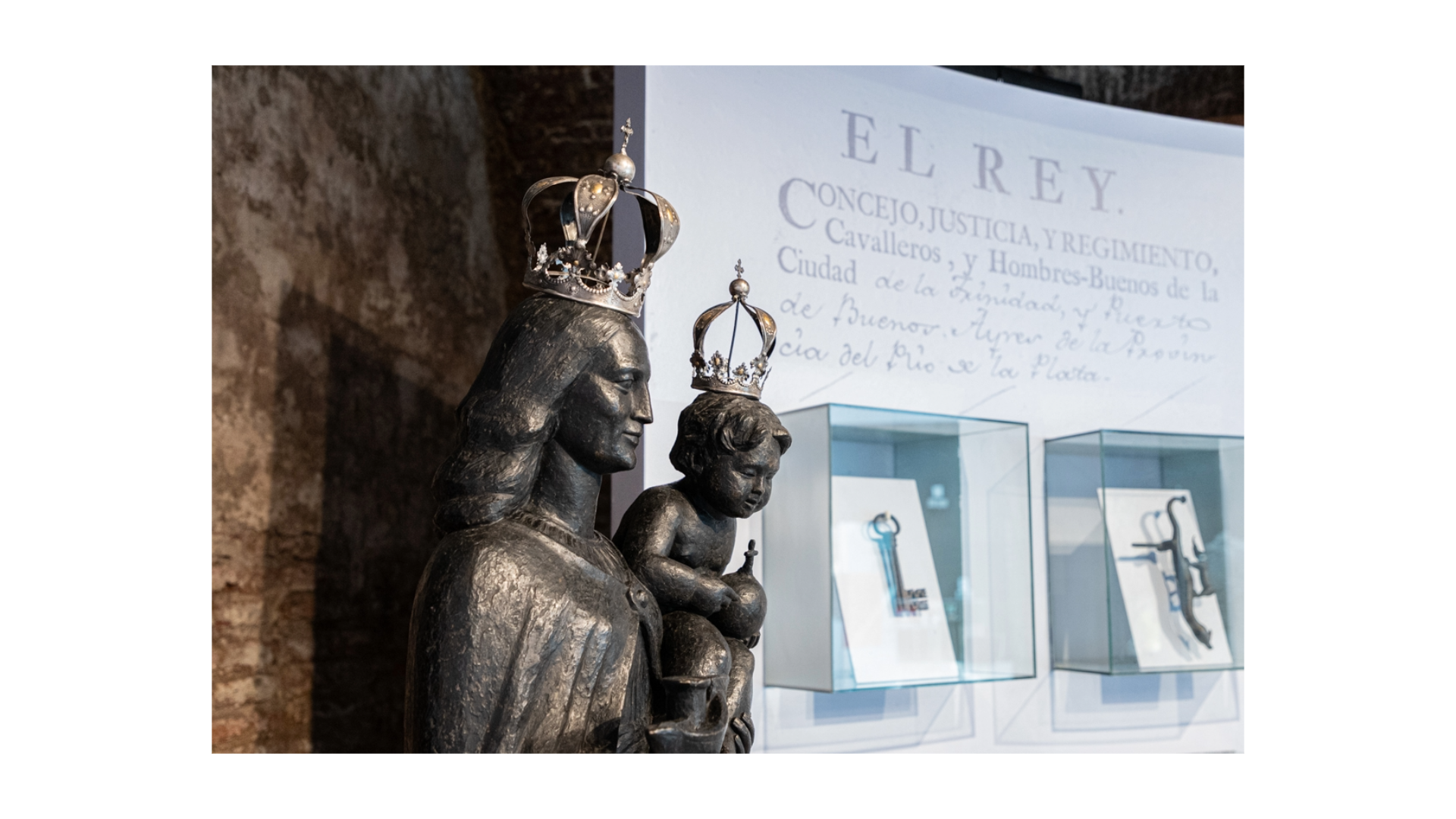 Detalle de la escultura de yeso pintada de negro de la Virgen del Buen Ayre. Lleva al niño en sus brazos. Ambas imágenes llevan una corona de plata sobre sus cabezas. De fondo hay un panel con una inscripción. También hay nichos de vidrio con piezas.