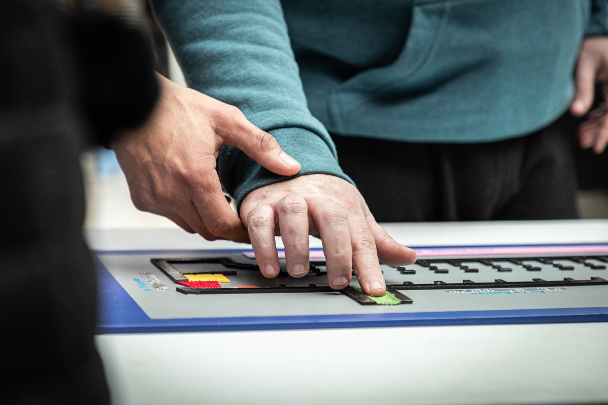 La mano de un hombre con buzo azul está tocando el plano háptico del Museo Casa Rosada asistido por otro hombre, del que solo vemos su mano, que viste buzo negro.