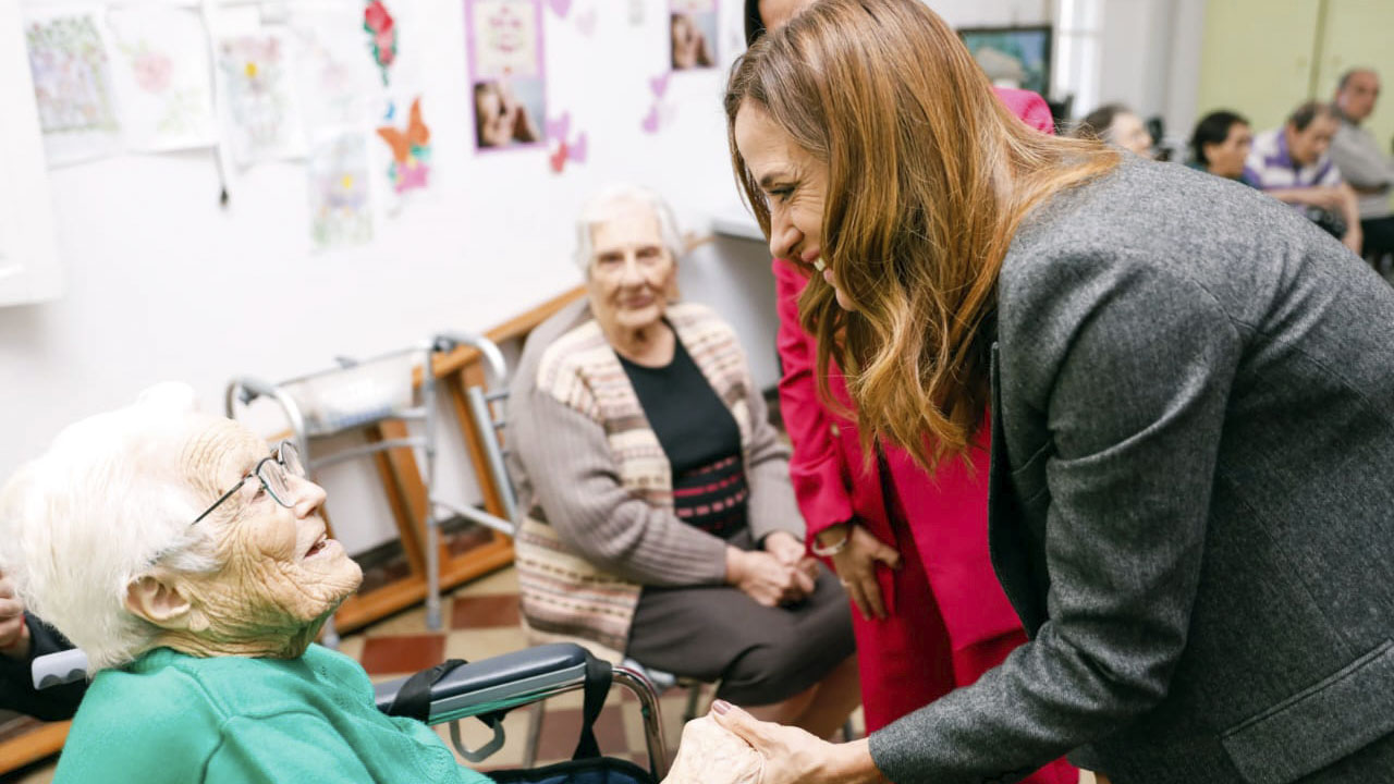 La ministra Victoria Tolosa Paz andole la mano y sonriendo a una mujer adulta mayor de pelo blanco, en silla de ruedas en el Hogar San José en Chacabuco.