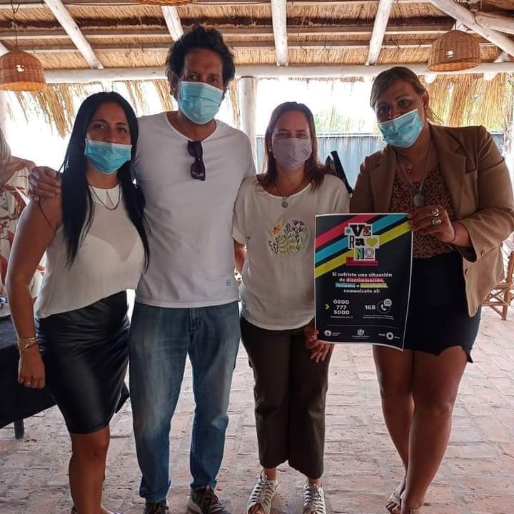 Junto a 2 personas del equipo de la delegación y la delegada Paula Saini, Victoria posa con un cartel de la campaña del INADI "Verano sin Discriminación".