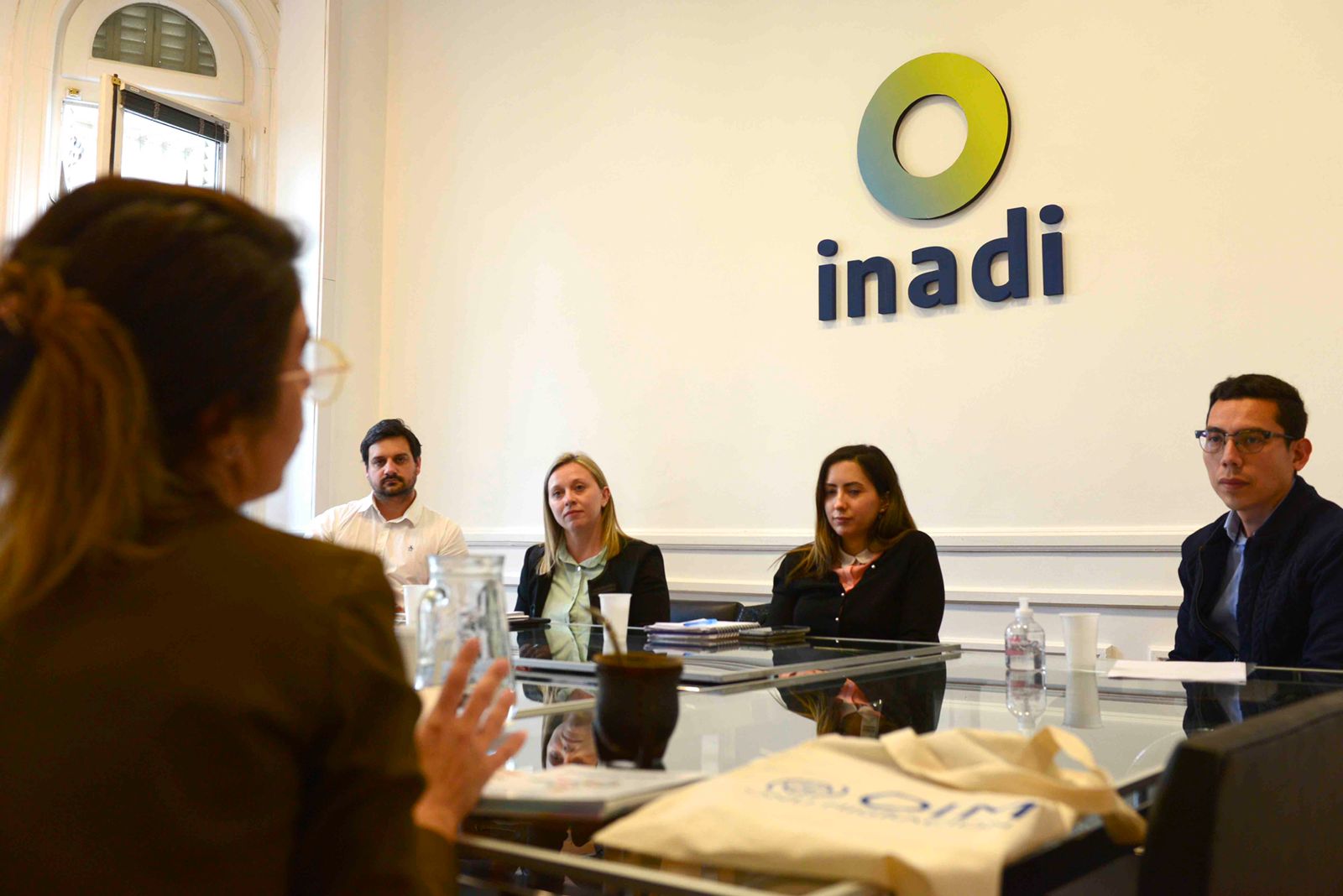 Participantes de la reunión conversan sentados en una mesa en la sala de reuniones con el logo de INADI en la pared de atrás