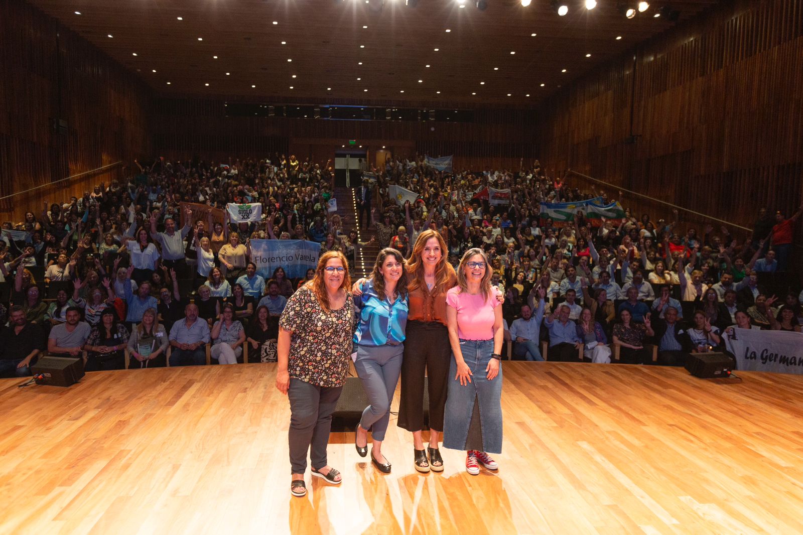 En el medio del escenario del auditorio "Ballena Azul" del Centro Cultural Kirchner, se encuentran paradas quienes expusieron el programa Participar en Comunidad: Victoria Tolosa Paz, Micaela Ferraro, Laura Berardo y Paola Rezano. De fondo se ve la multitud que participó del encuentro.