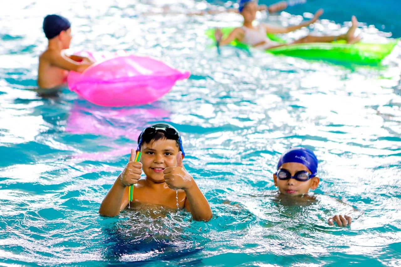 Niños jugando en la pileta de natación con inflables y accesorios de natación, uno de ellos saludando a la cámara.