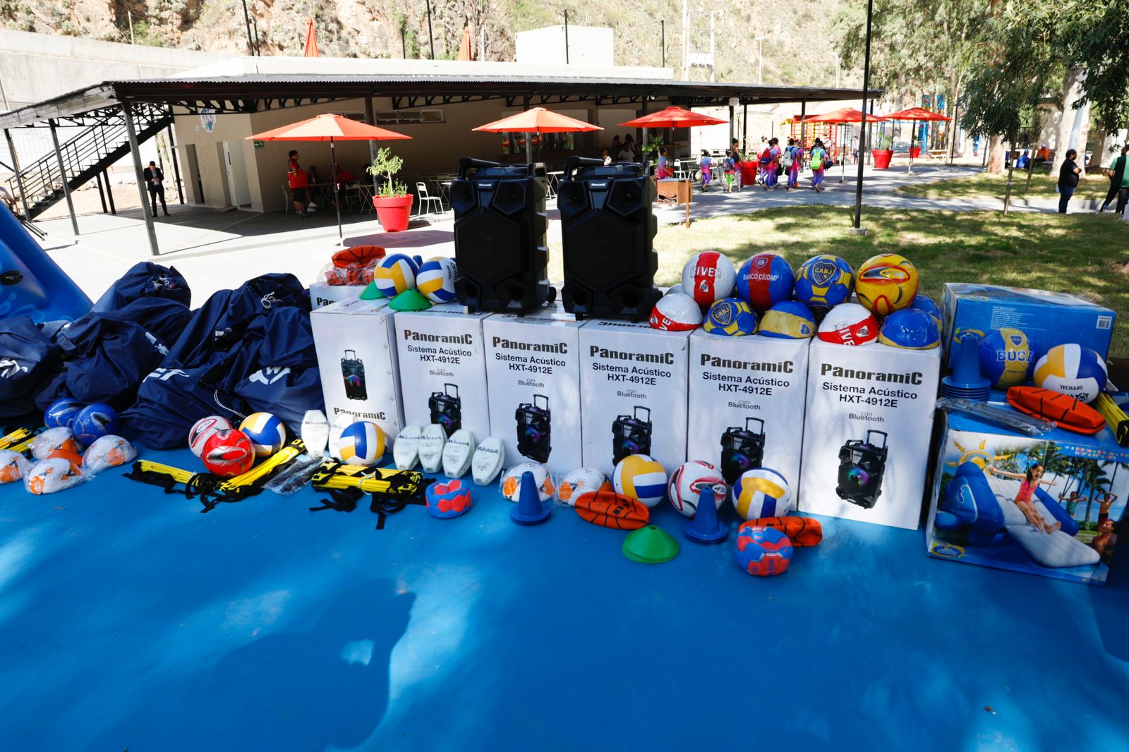 Serie de materiales deportivos, pelotas, conos, inflables nuevos en sus cajas colocadas una al lado de la otra sobre un paño azul.