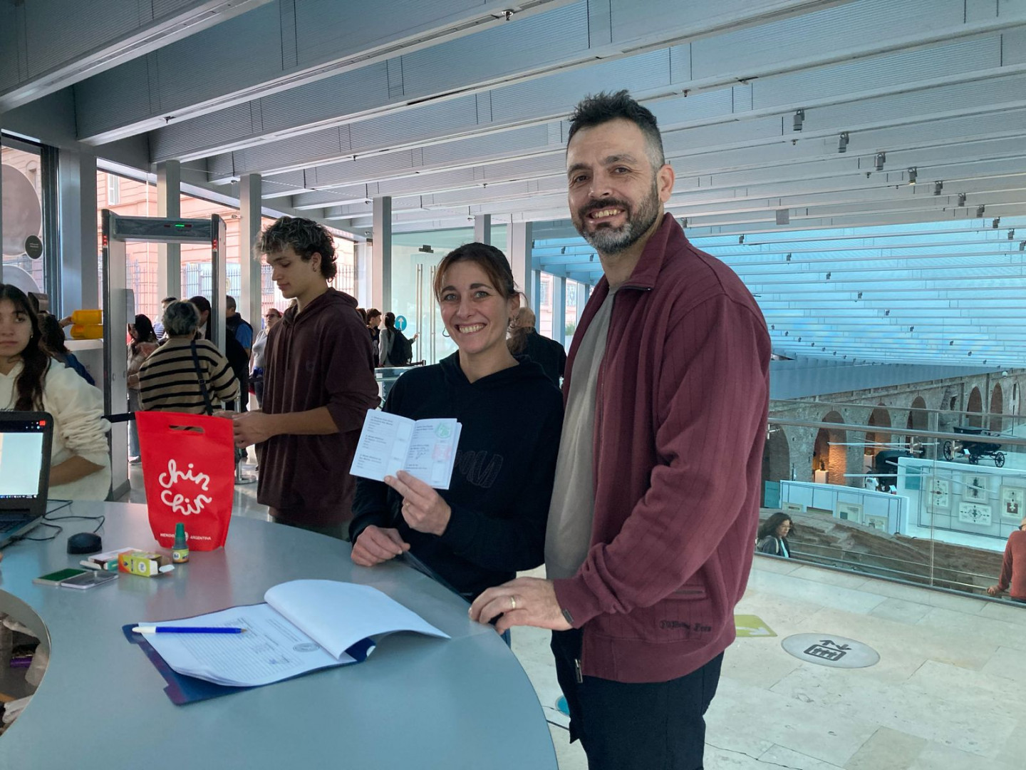 Dos personas miran a cámara. La mujer se encuentra hacia la izquierda y lleva un sweater de color negro. En su mano lleva una copia del Pasaporte Nacional Sanmartiniano. A su derecha un hombre que viste campera de color rojo la mira sonriente.
